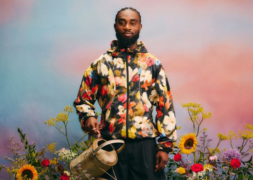 Kofi Stone wearing a floral jacket surrounded by flowers holding a watering canx